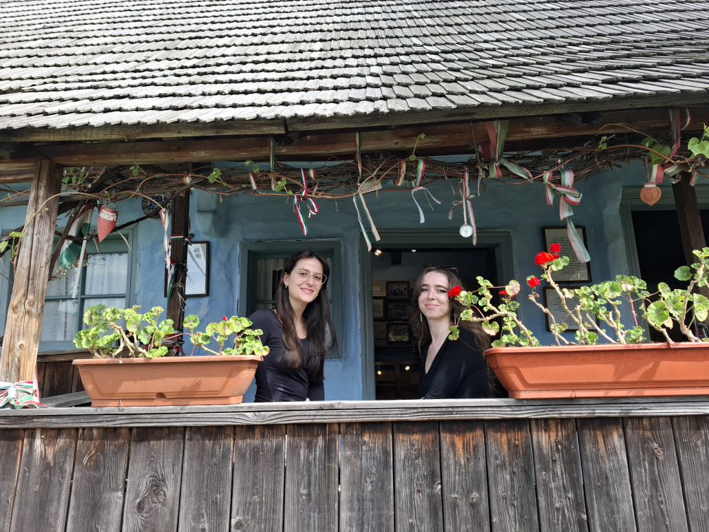 Two scholarship holders on the porch of a traditional Hungarian house during a cultural visit, surrounded by flowers and folk-inspired decorations.